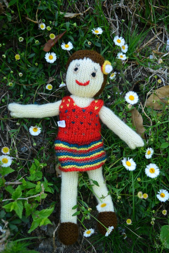 Knitted soft toy of a small girl in a red from lying o a bed of flowers